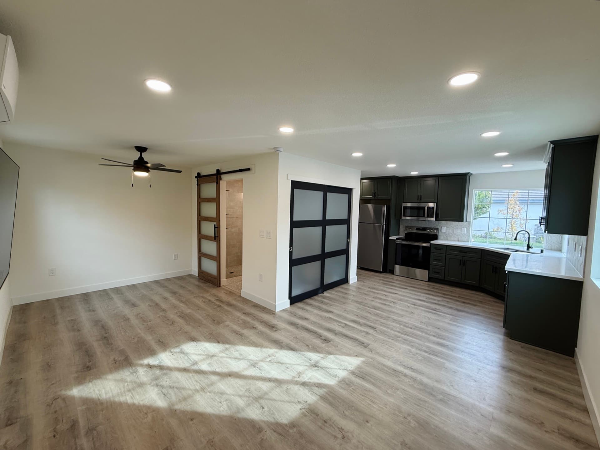 Open-plan converted garage ADU with full kitchen, dark green shaker cabinets, stainless appliances, sliding barn door and luxury vinyl plank flooring in a Sacramento garage-to-ADU conversion